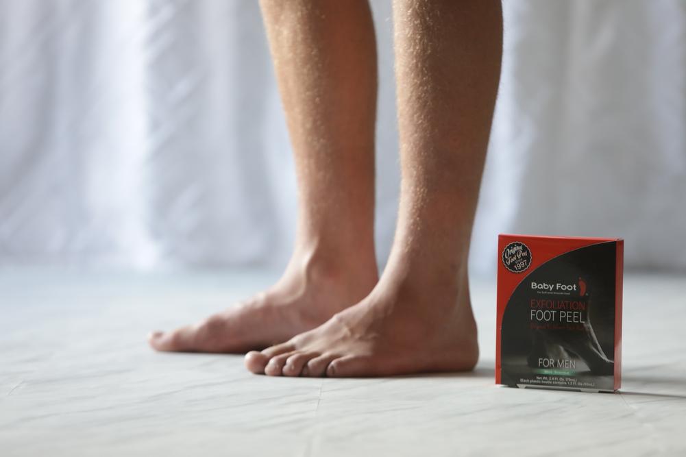 A man's feet standing next to a package of Baby Foot 1-Hour Men's Foot Treatment