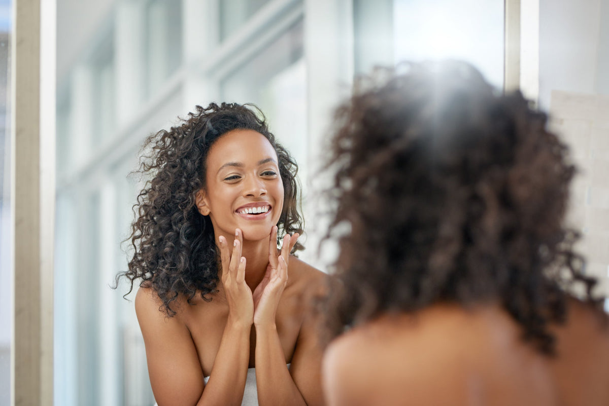A woman wearing a towel around her torso, smiling while looking at herself in the mirror.