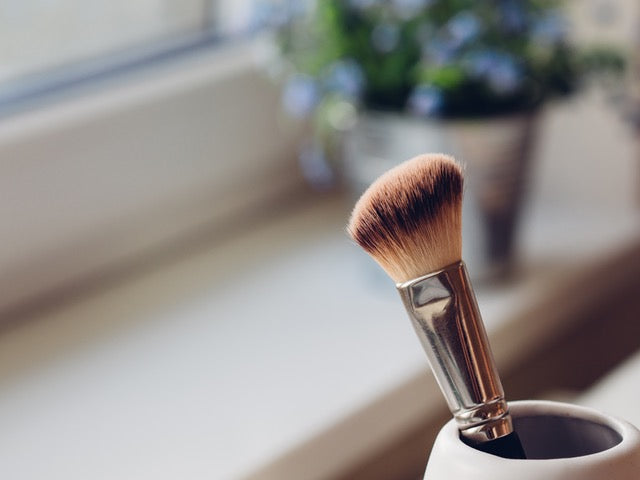 a large makeup brush positioned bristle end-up, in a white ceramic jar. in the background there is a windowsill and a potted plant