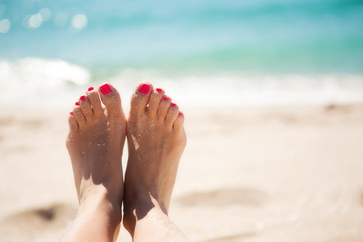 A woman holding up her sand-covered feet up with the ocean in the background.