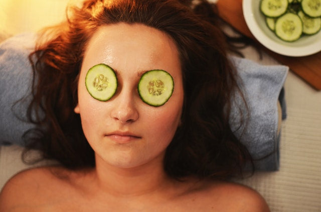 a woman lays with cucumber slices over her eyes, her head is rested on a towel, next to a plate of cucumber slices